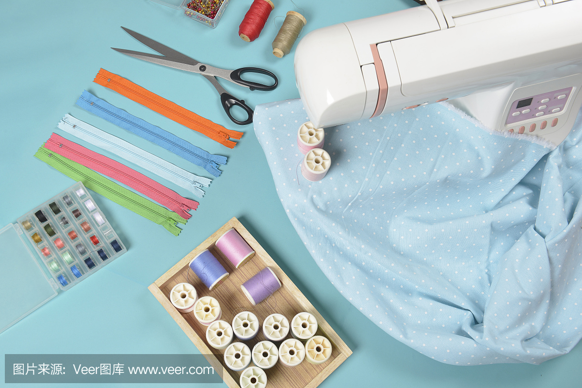 缝纫机上的布料夹在剪刀、衬衫纽扣、拉链、别针和蓝色背景上缝制的彩色线卷之间。